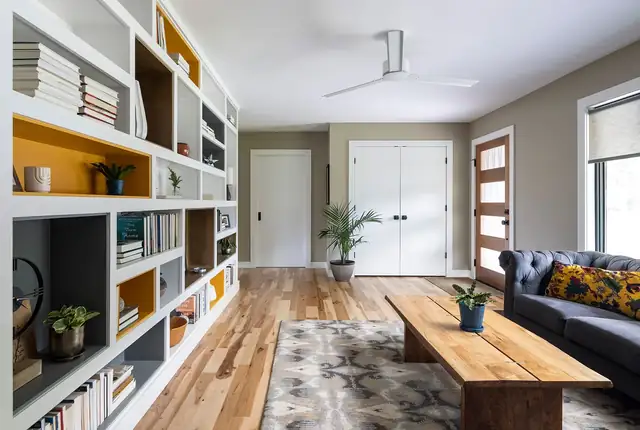 Remodeled living room with built-in white bookshelves with yellow backs, wide-plank wood floor and large windows