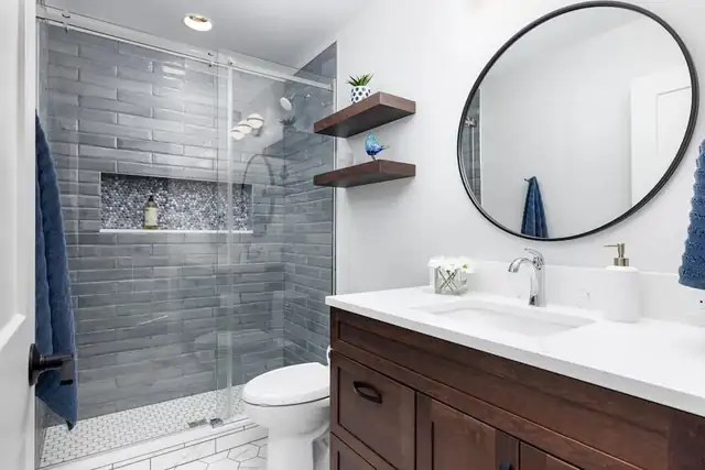 Bathroom with gray glass-tile walk-in shower, tiled niche, floating wood shelves and round black mirror