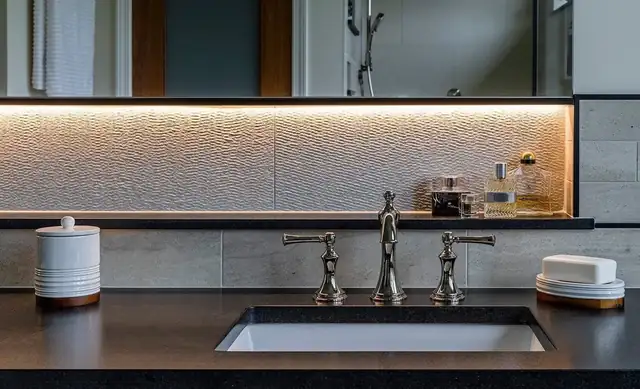 Bathroom vanity with a backlit mirror, textured tile wall, dark countertop and widespread faucet in an Ann Arbor home